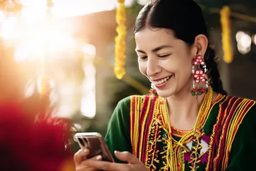 Uma mulher alegre em roupas tradicionais usando seu smartphone durante um momento festivo, mostrando como o app BB22 se encaixa facilmente no dia a dia.